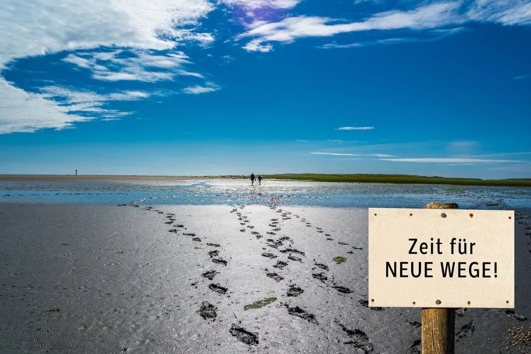 Schild „Zeit für neue Wege!“ im Watt unter blauem Himmel